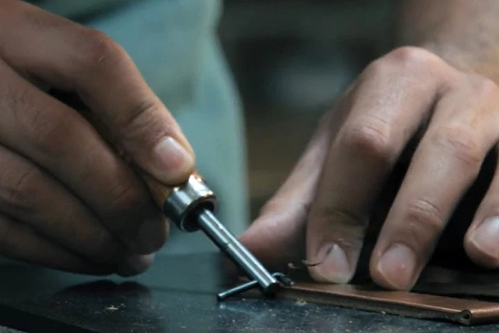 Hände führen ein Metallwerkzeug entlang der Kante von braunem Leder